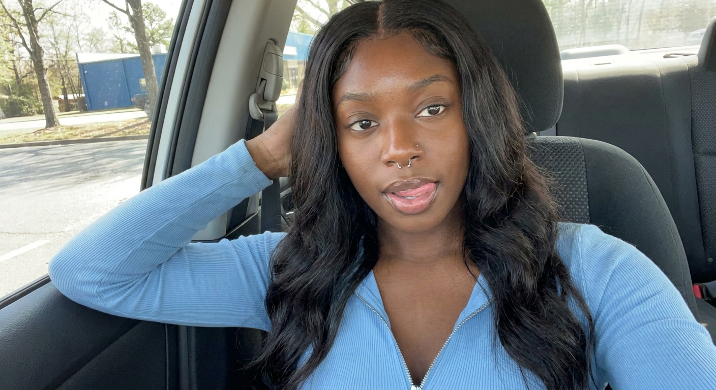 **Ultra-Detailed Description:**

The image is a candid, close-up selfie of a young Black woman seated in the back seat of a car. She has rich, deep brown skin with a smooth, natural glow and is looking directly at the camera with a relaxed, slightly playful expression. Her head is tilted gently to her right, and her lips are parted with her tongue lightly touching her lower lip in a casual, confident pose. She has a silver septum nose ring and an additional small silver stud on her left nostril.

Her hair is long, jet-black, and styled in sleek, voluminous waves with a middle part. The hair cascades luxuriously over her shoulders and chest, with soft layers and subtle body that frame her face beautifully. A few strands fall naturally across her forehead and cheek.

She is wearing a light blue (minty/aqua-toned) long-sleeved ribbed zip-up top. The top has a partially unzipped front that creates a flattering V-neckline, revealing her décolletage. The fabric appears soft and stretchy with a fine waffle/ribbed texture. Her left hand is raised behind her head, fingers loosely gripping the headrest or the back of the seat, showcasing her manicured nails.

The setting is the interior of a vehicle, likely an SUV or larger car, with dark gray fabric seats visible behind her. Through the car window on the left side of the frame, daylight reveals an outdoor scene with green trees and what appears to be a parking lot or roadside area, including a glimpse of a blue structure in the distance. The natural daylight coming through the window illuminates her from the side, creating soft highlights on her skin and hair while keeping the overall mood intimate and casual.

The composition has a natural, everyday vibe typical of a personal selfie taken while riding or parked in a car, with the focus sharply on her face, hair, and upper body. The color palette is dominated by the cool tones of her top, the dark richness of her hair, and the neutral grays of the car interior, contrasted by the greenery outside.